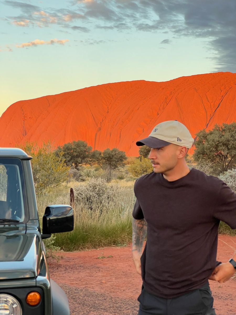 Billy in a cap and dark T-shirt, standing in front of Uluru glowing red at sunset